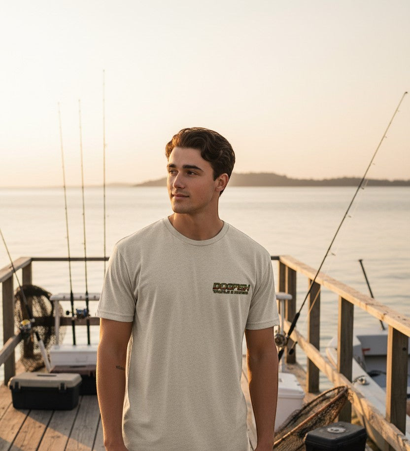 Man standing on a dock with fishing rods, wearing a t-shirt with a logo, during sunset.