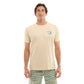 A man modeling a light beige crewneck t-shirt with a small blue wave graphic on the left chest, paired with green and beige patterned swim trunks, against a solid white background.
