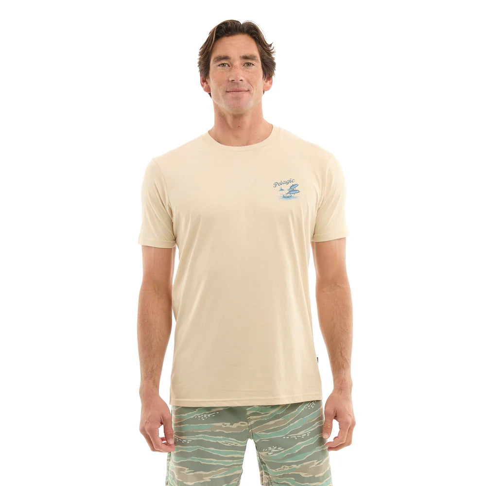 A man modeling a light beige crewneck t-shirt with a small blue wave graphic on the left chest, paired with green and beige patterned swim trunks, against a solid white background.
