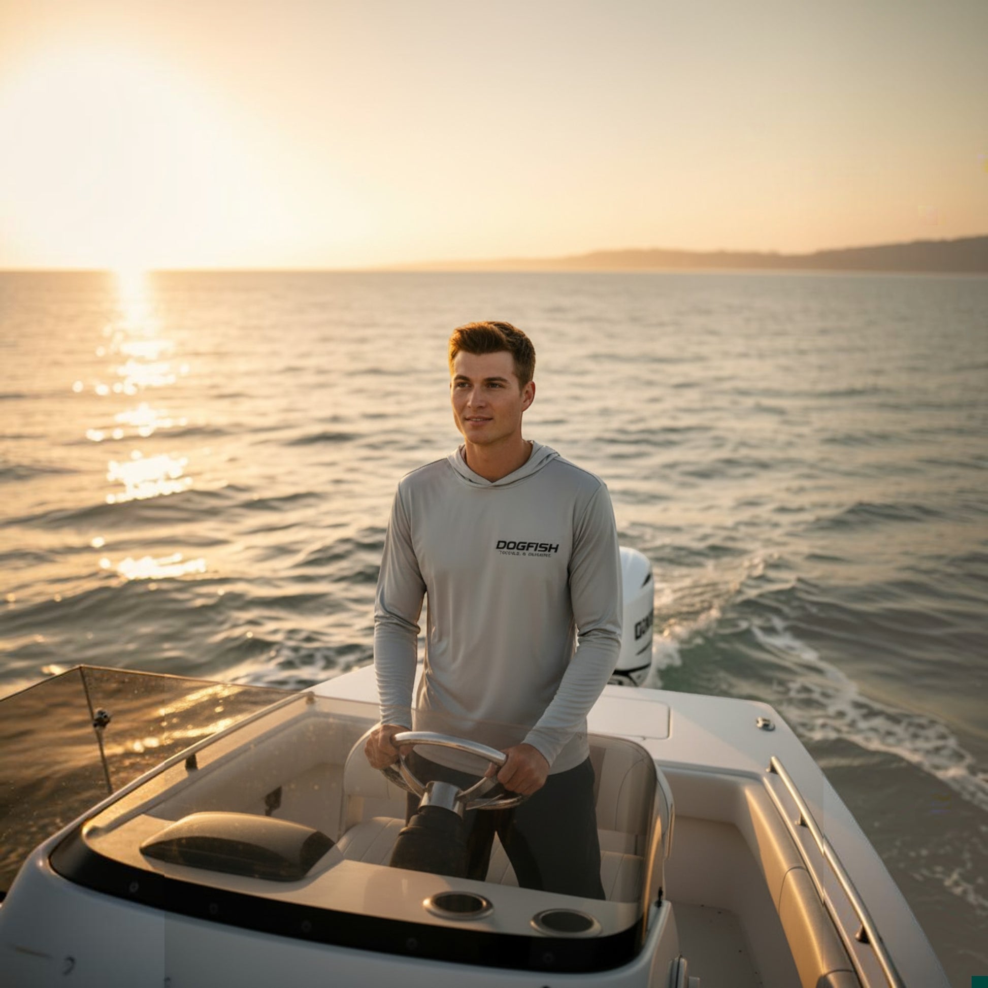 Man on a boat wearing a gray long-sleeve shirt with a logo, steering the boat at sunset.