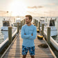 Dogfish Tackle & Marine light blue microfiber long sleeve shirt with logo, worn by a man on a dock with boats.