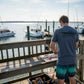 Dogfish Tackle & Marine heathered navy blue fishing boat graphic t-shirt worn by a man cleaning fish on a dock.