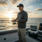 Dogfish Tackle & Marine logo trucker hat with camouflage pattern and mesh back, worn by a man fishing on a boat at sunset.