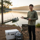 Dogfish Tackle & Marine olive green long sleeve microfiber shirt with crew neck and black logo. Model stands by a lake with fishing gear.