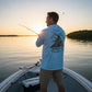 Man wearing a light blue Dogfish Tackle & Marine long sleeve performance hoodie while fishing on a boat at sunset.