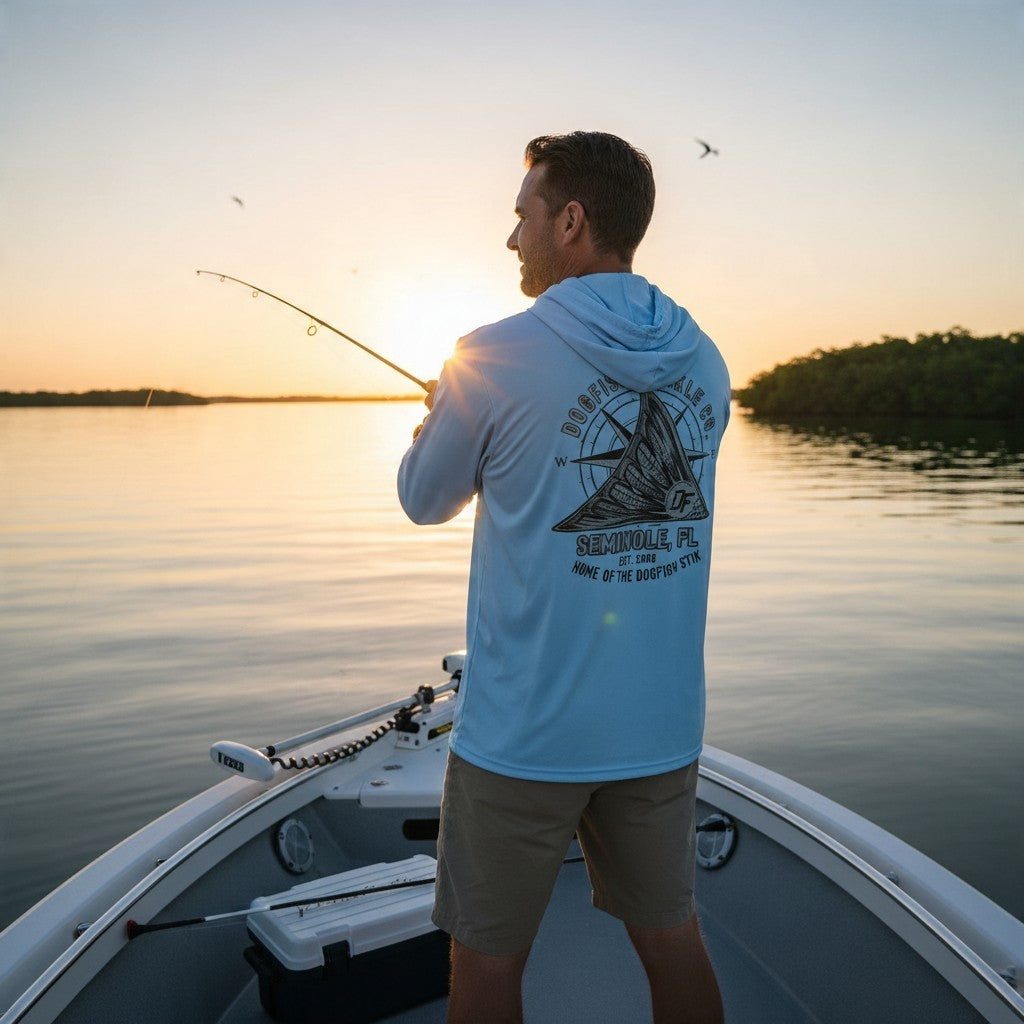 Man wearing a light blue Dogfish Tackle & Marine long sleeve performance hoodie while fishing on a boat at sunset.