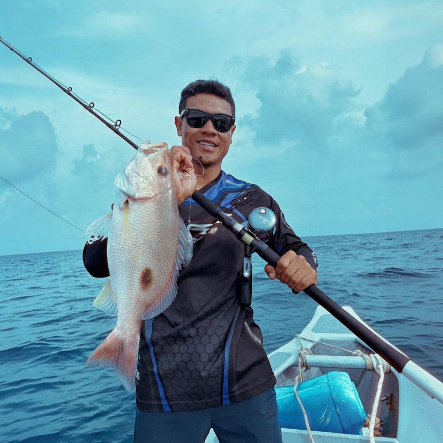 Dogfish Tackle & Marine man holding a large fish while fishing from a boat, wearing a black and blue long-sleeve shirt.